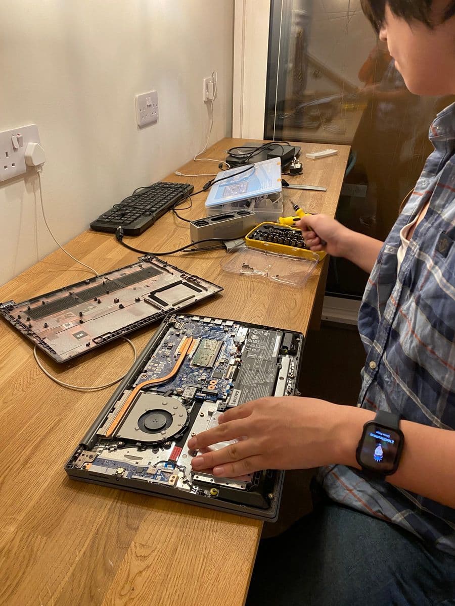 Hands working on the inside of an opened laptop on a desk, with a screwdriver and a tray of parts